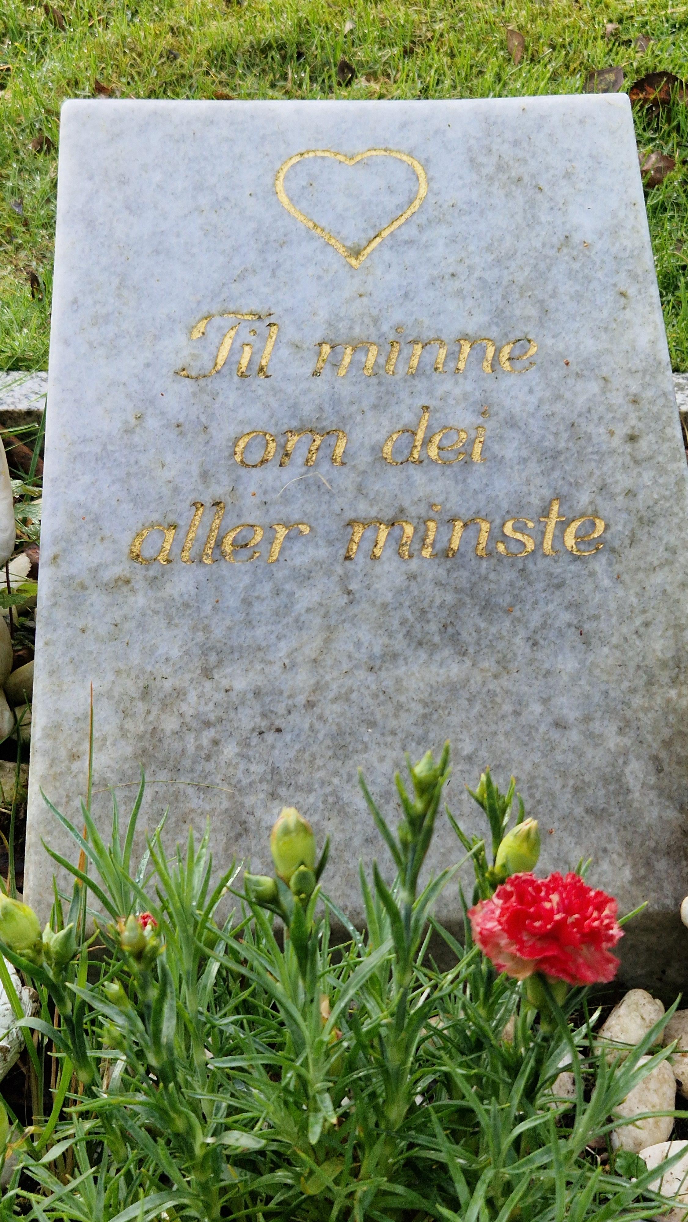 Bilde viser eit lite grått gravminne som står på skrått bakover med gull tekst som viser eit hjerte og tekst som står, til minne om dei aller minste. Vi ser også blomster i blomsterbeddet.